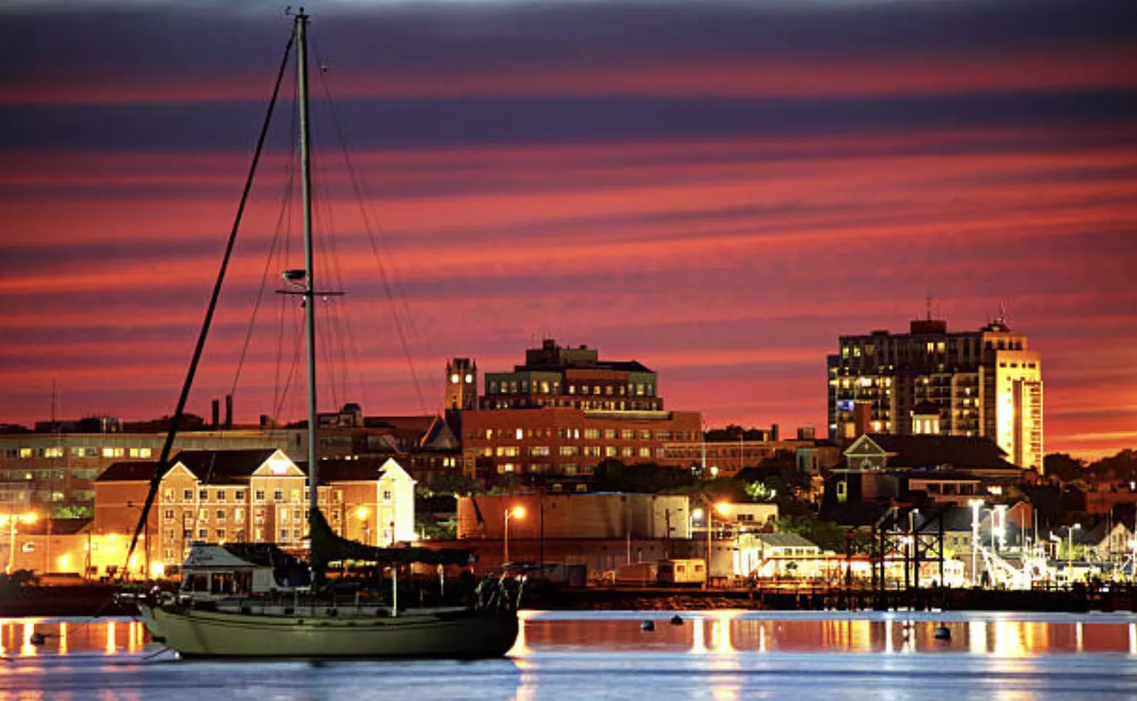 New Bedford, Massachusetts skyline across the harbor at sunset, downtown brick office buildings and a clock tower lit under a red sky, with a moored sailboat in the foreground