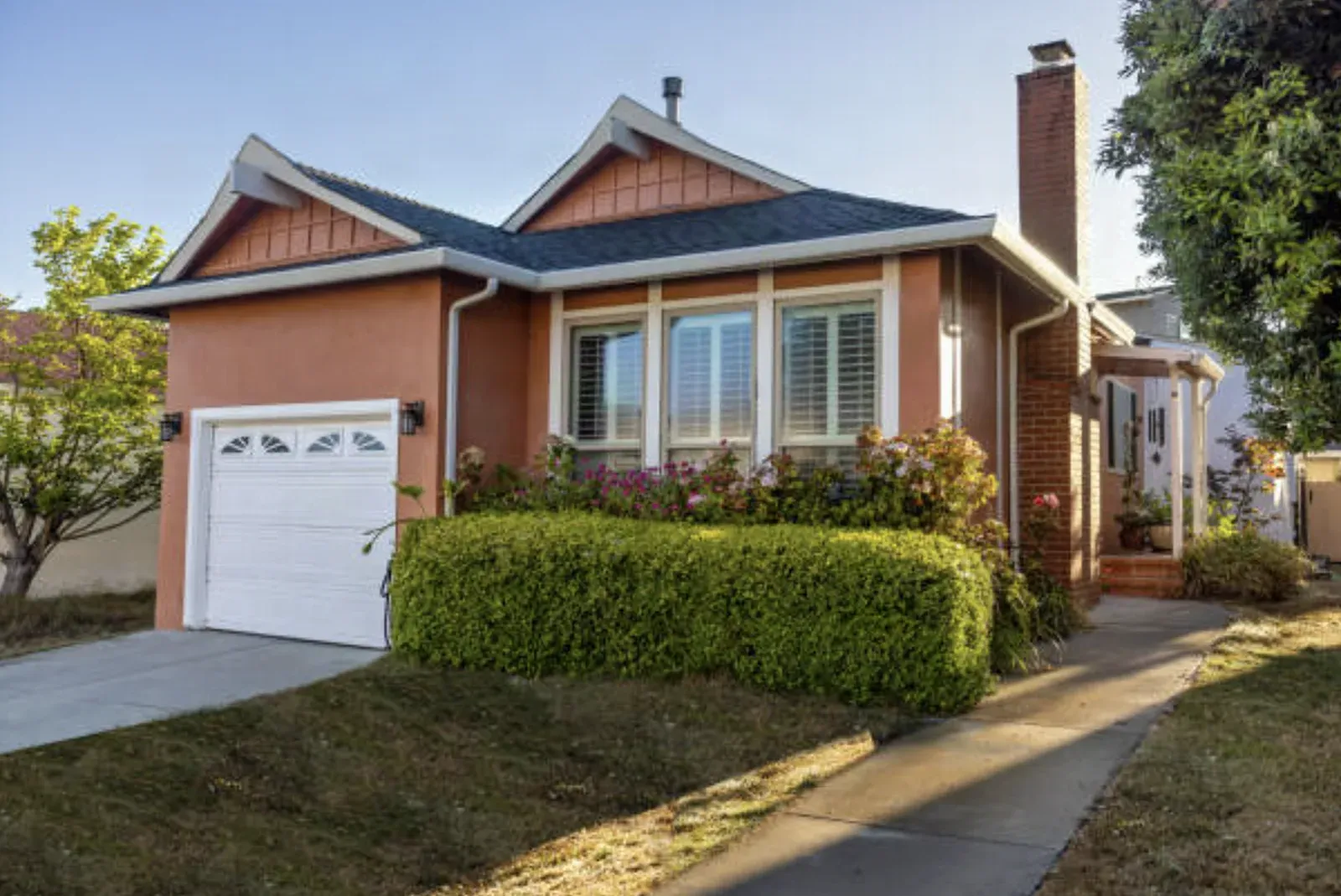 Single-story rental house with a stucco exterior, attached one-car garage, bay of shuttered front windows, and a trimmed hedge along the front walk