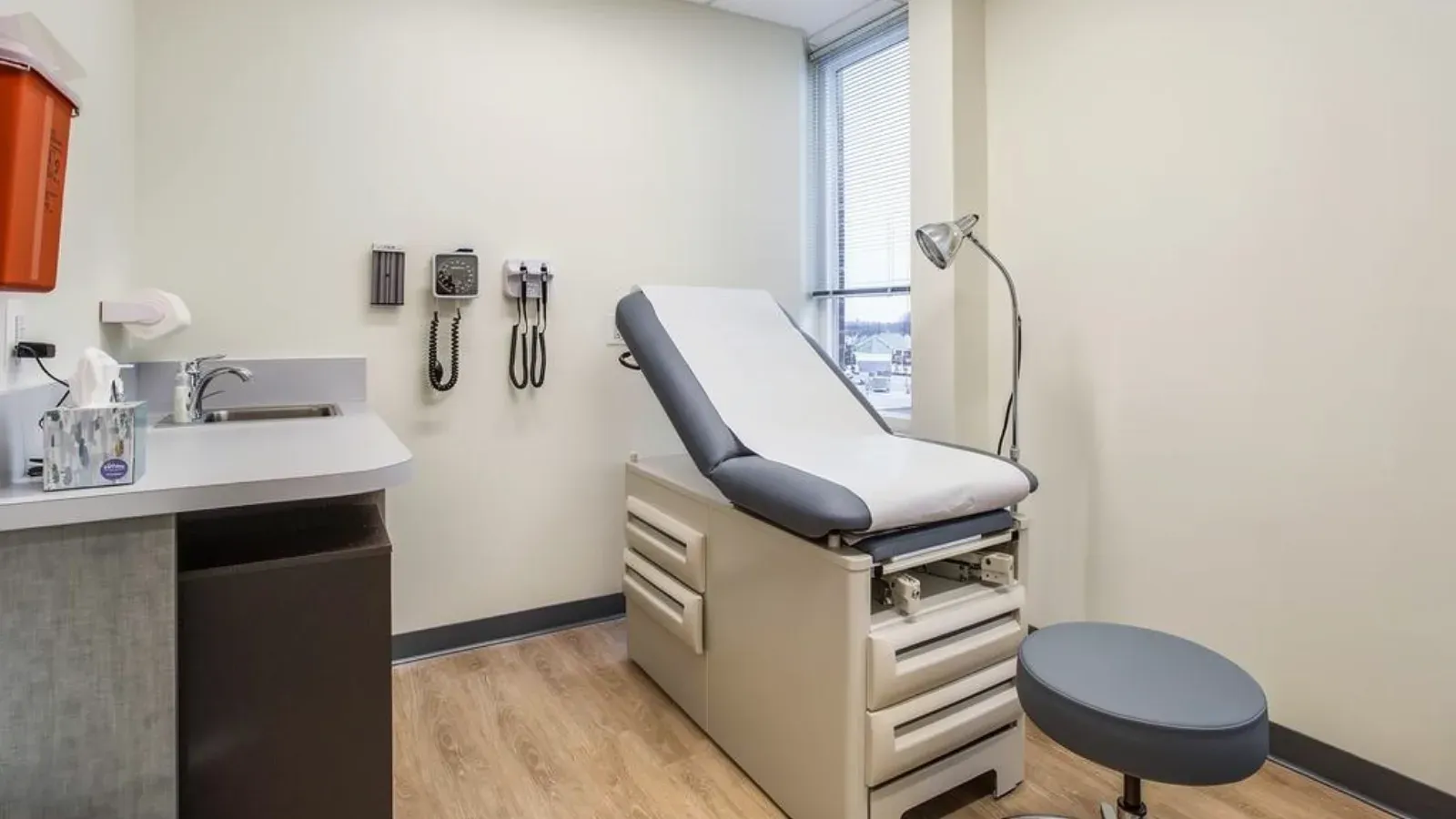 A medical exam room with an exam table against a broad plain wall, a sink counter and wall-mounted diagnostic instruments