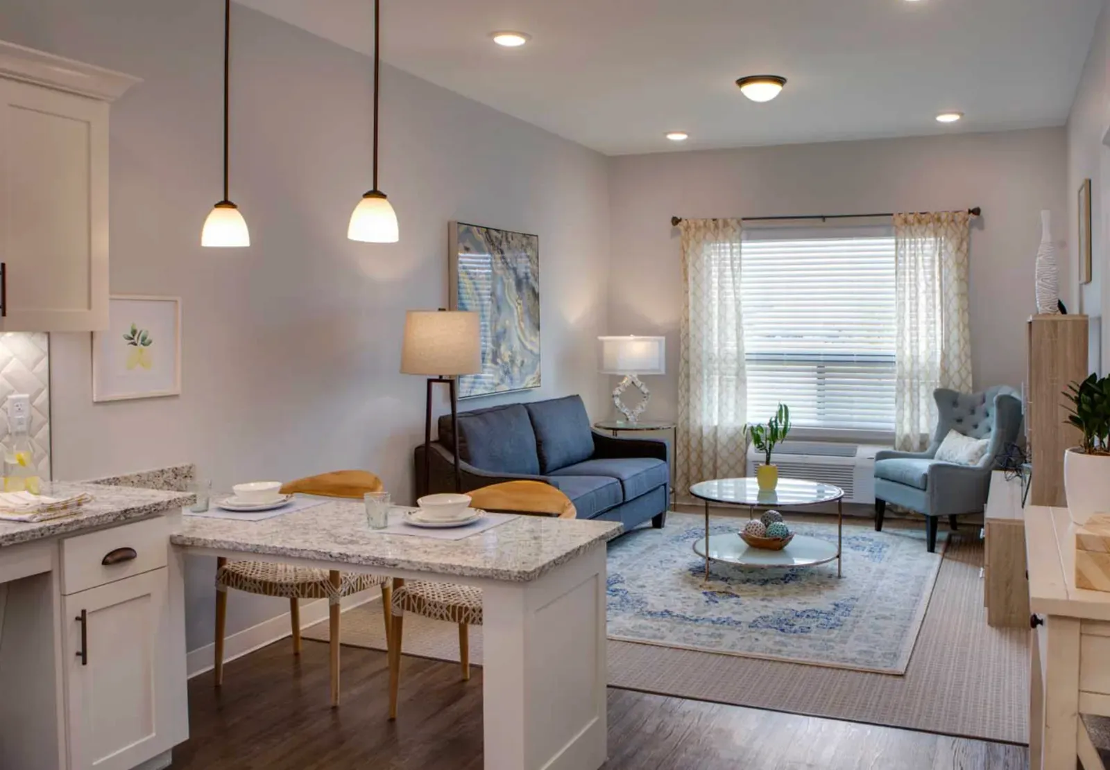 Senior-living apartment unit with finished painted drywall, recessed and pendant lighting, an open kitchenette and dining table beside a living area with a sofa, armchair, and curtained window